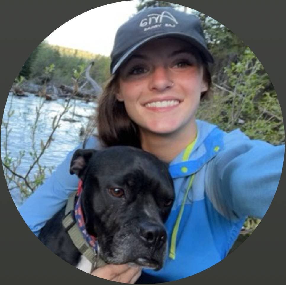 A smiling woman in a cap holds a black dog beside a river while taking a selfie outdoors.