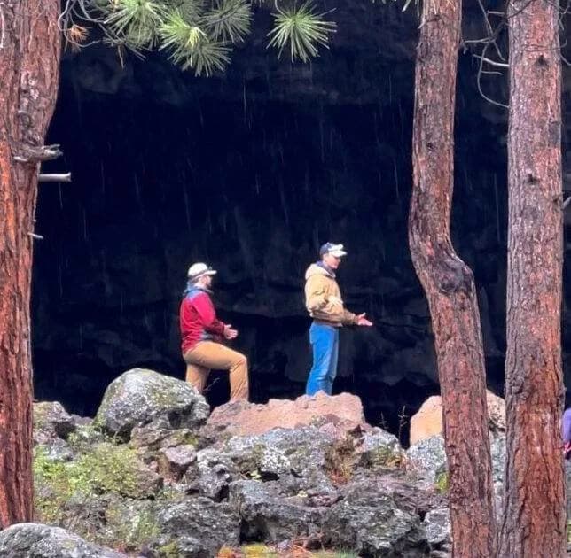Two people sit on rocks among tall trees in a forest setting.