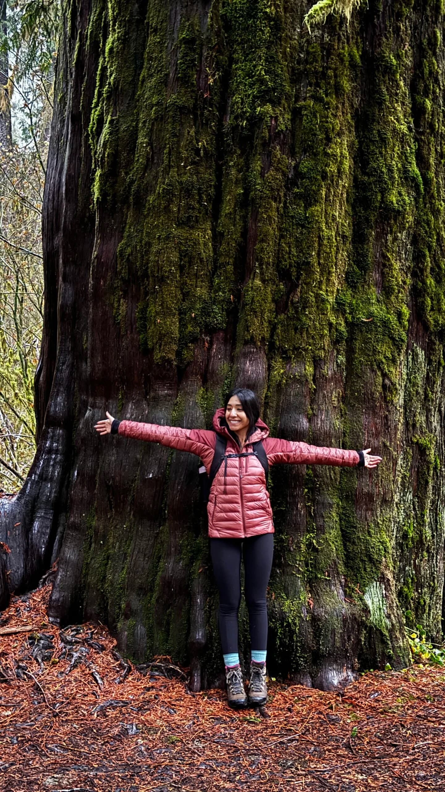 A smiling woman stands in front of a moss-covered tree with her arms outstretched.