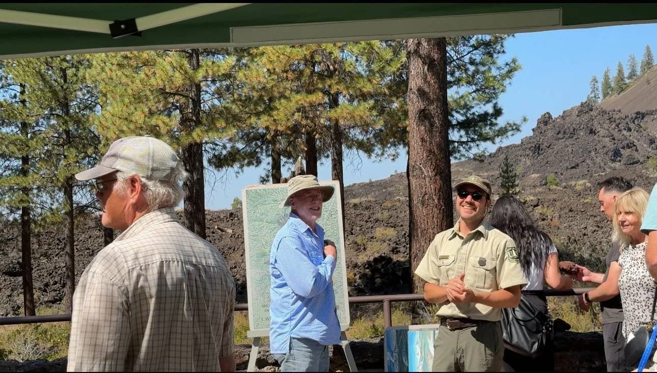 Group of people outdoors, some wearing hats and sunglasses, standing near a sign with a forested background and rocky hillside.