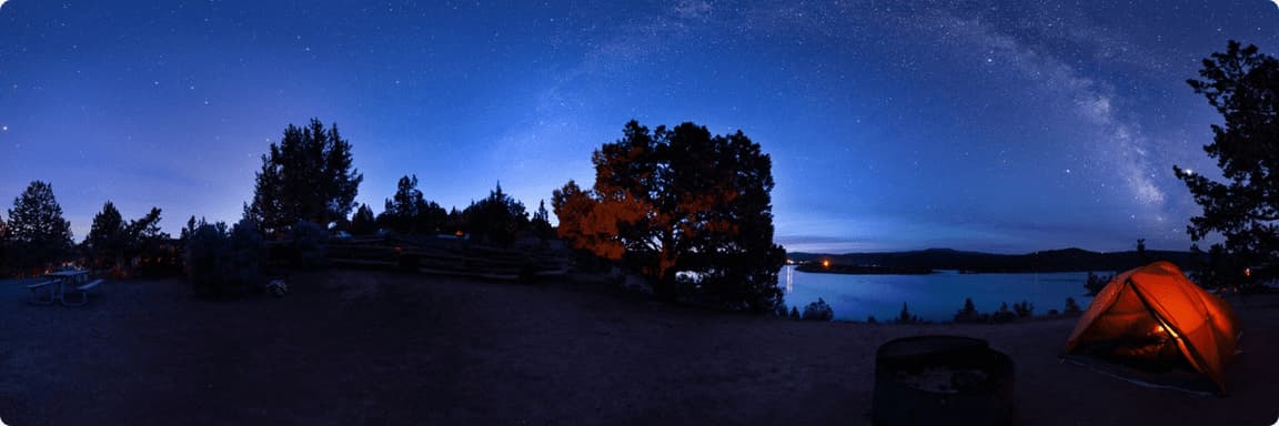 Camping tent glowing under starry night sky near lake with Milky Way visible