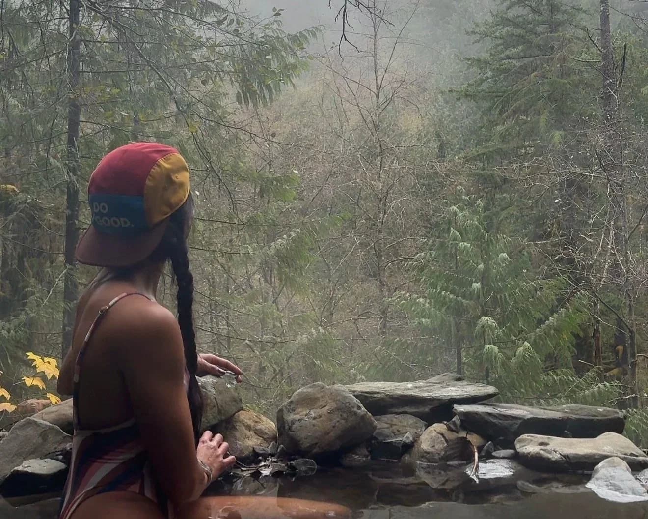 Person in swimsuit and cap relaxing in forest hot spring surrounded by misty trees.