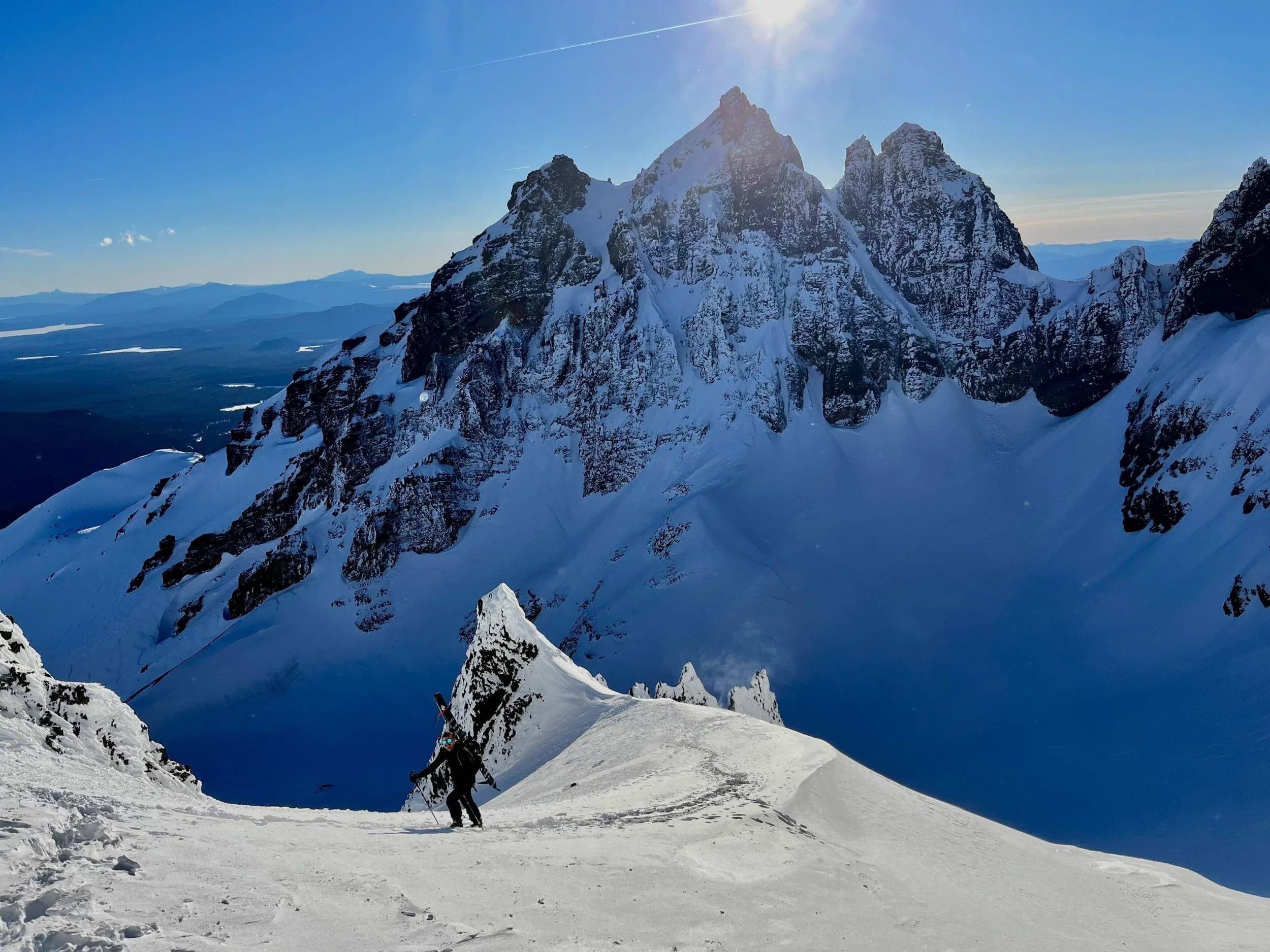 Skier on snowy mountain ridge with jagged peaks under blue sky