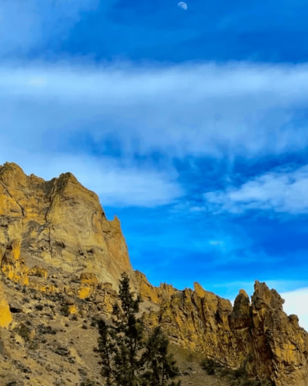Rugged golden cliffs under blue sky with moon, ideal for hiking and climbing adventures.