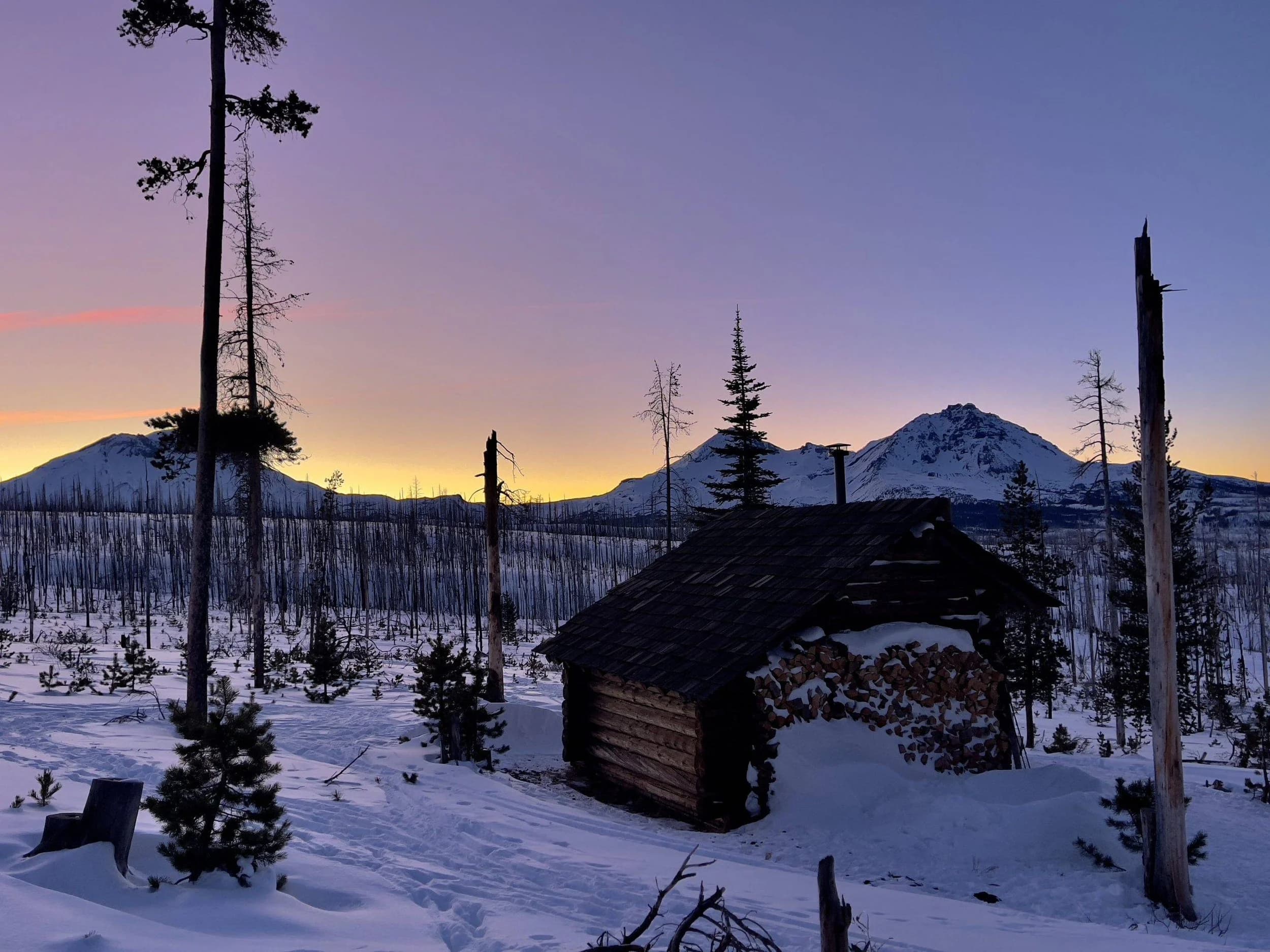 Snowy cabin at dusk with mountains and trees, ideal for winter adventures.