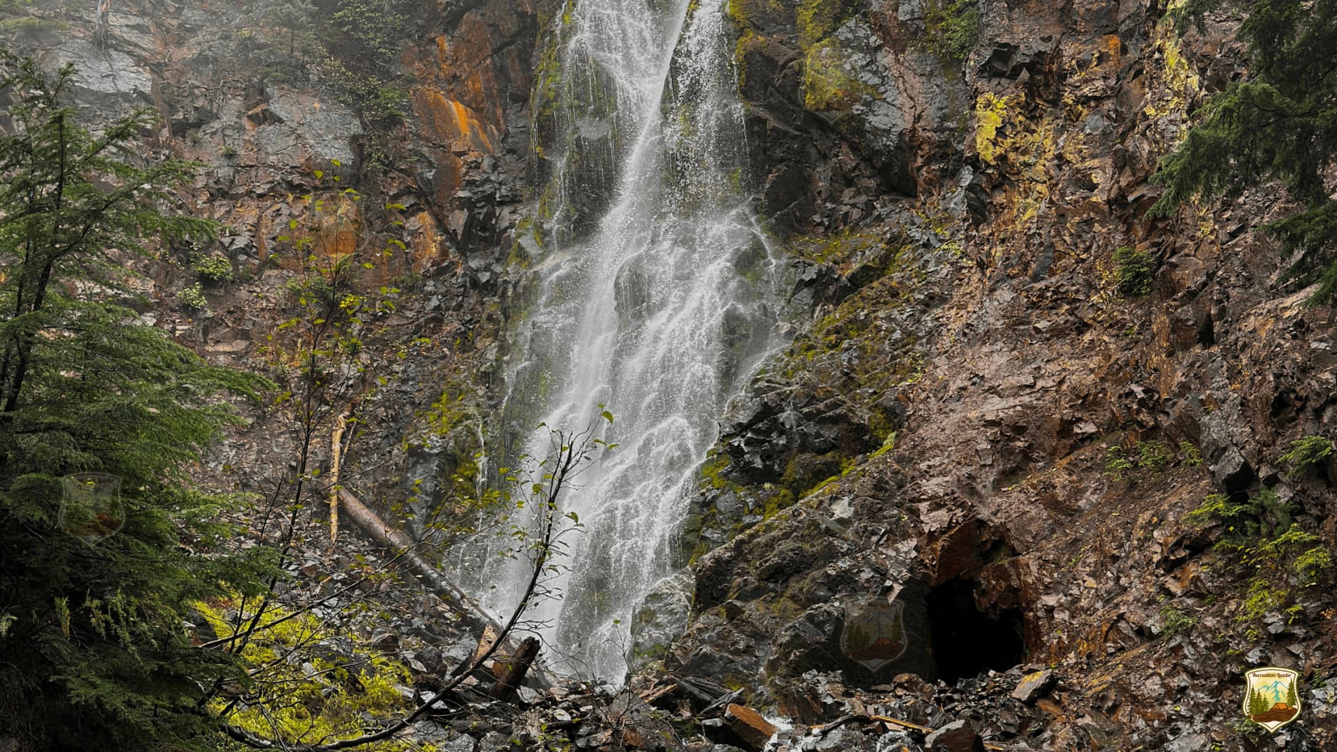 Waterfall cascading down rocky cliff with moss and trees, near a cave entrance.