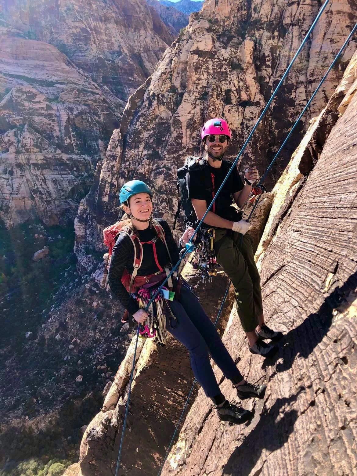 Two climbers smiling on a cliff face with ropes and gear, mountains behind.