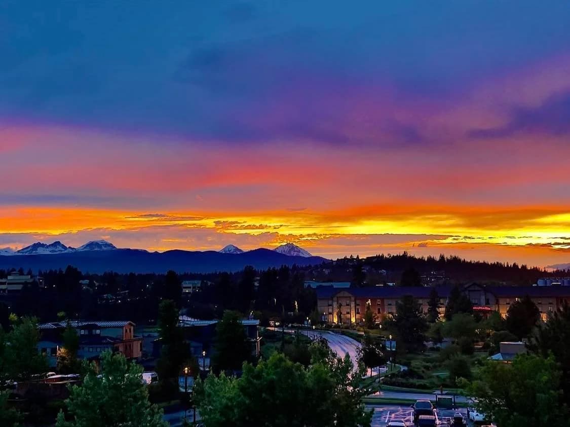 Sunset over mountains with town below, trees and buildings lit up at dusk.