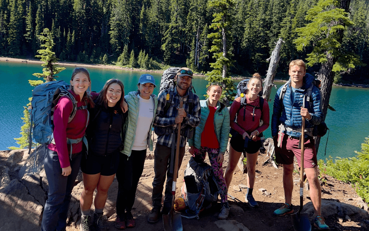 A group of six backpackers at a lake in the Three Sisters Wilderness volunteering; some hold shovels.