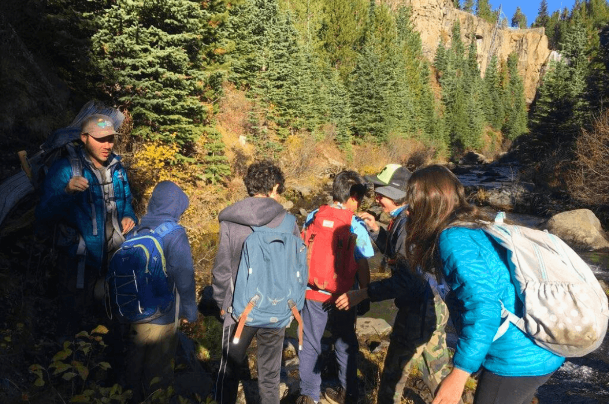 A group of seven hikers, including males and females, standing on rocks near a river in a forested area during fall, with evergreen trees and rocky cliffs in the background.