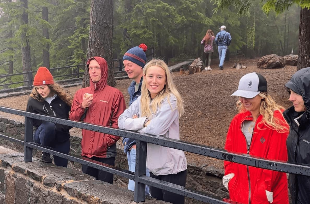 Five people standing behind a metal railing in a forested park, wearing jackets and hats, with two more people walking on a trail in the background.