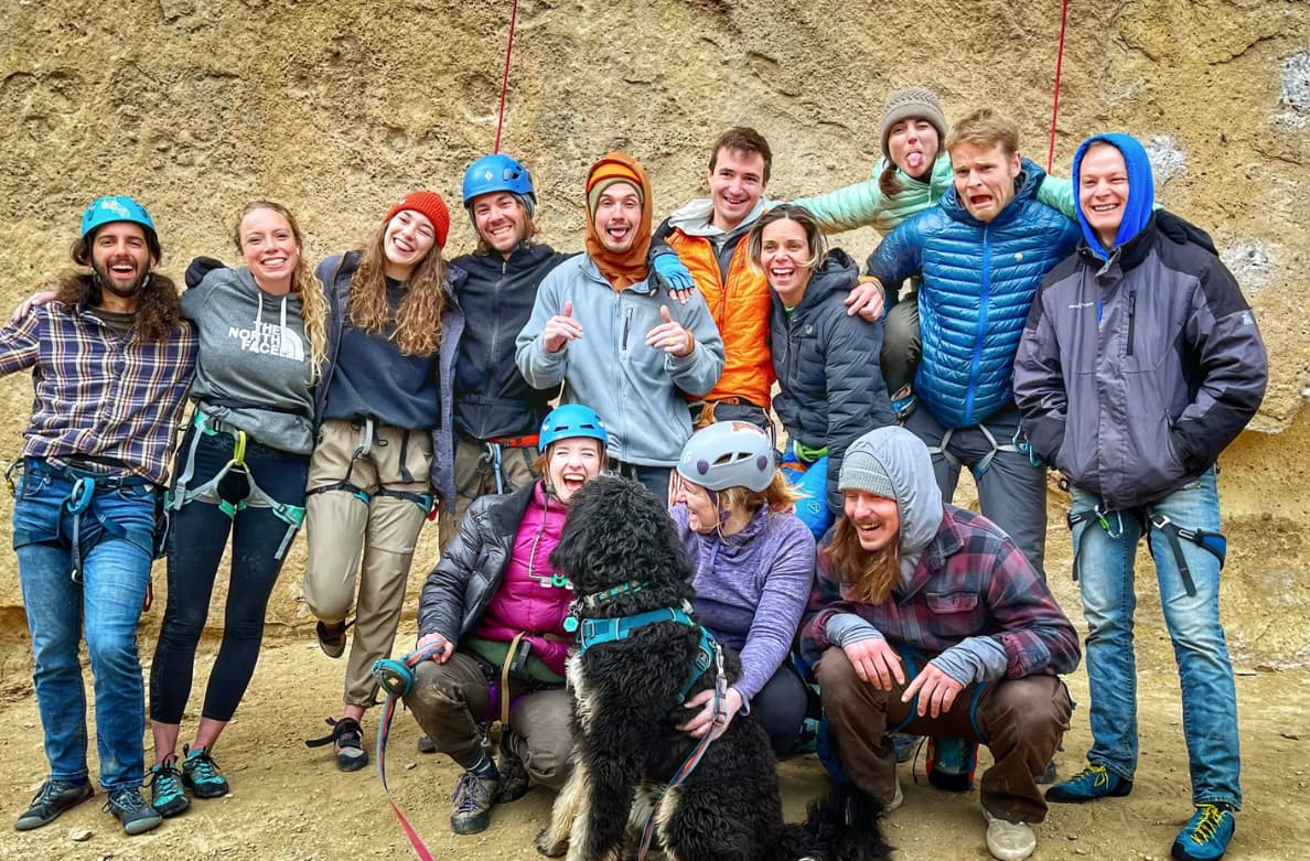 Group of people in outdoor rock climbing gear, standing and sitting against a rocky wall, smiling and posing for the photo, with a black dog among them.